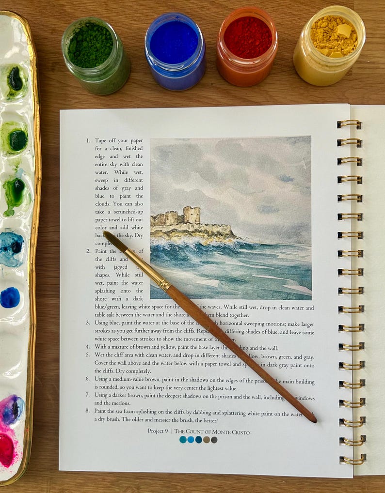 Puede incluir: Un diario de arte abierto con una acuarela de una escena costera, un pincel y peque&ntilde;os botes de pintura. El texto de la p&aacute;gina dice "Project 9 | THE COUNT OF MONTE CRISTO".