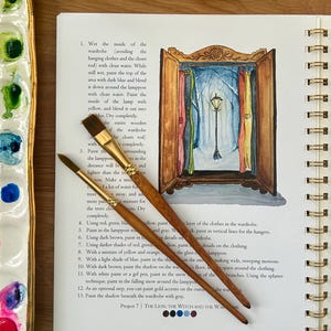 Puede incluir: Un libro abierto con ilustraciones en acuarela e instrucciones para un proyecto de pintura. Dos pinceles descansan sobre la p&aacute;gina, junto a una paleta de pinturas. El proyecto representa un armario con una farola en una escena nevada.