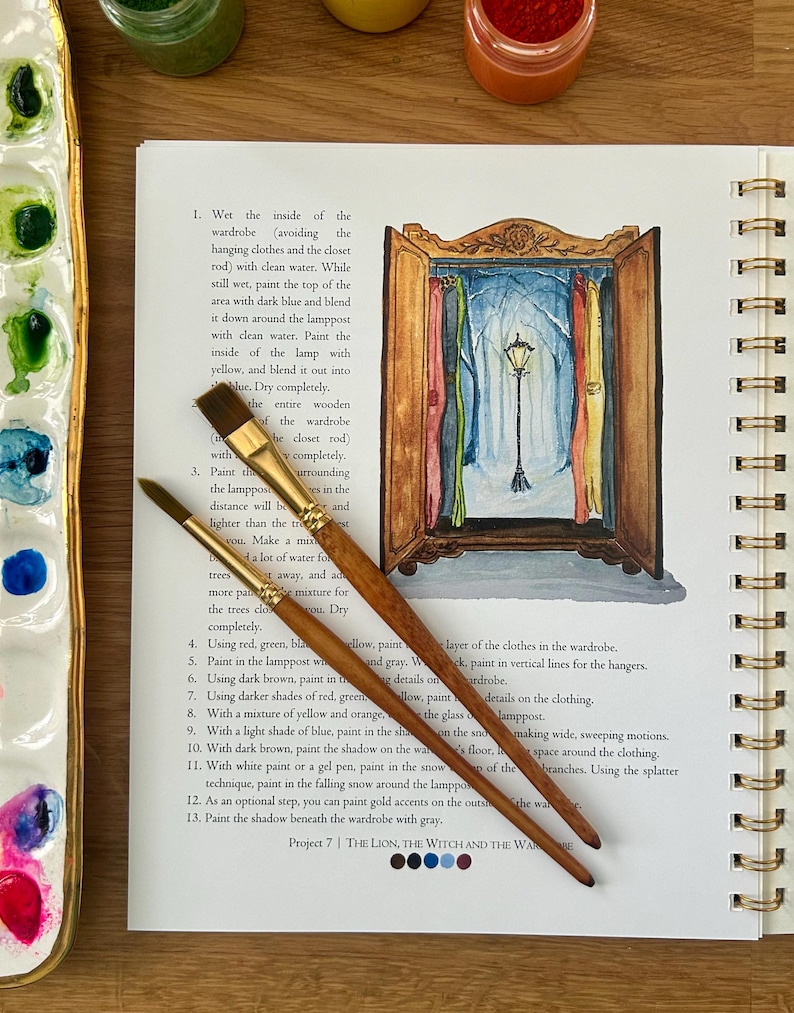 Puede incluir: Un libro abierto con ilustraciones en acuarela e instrucciones para un proyecto de pintura. Dos pinceles descansan sobre la p&aacute;gina, junto a una paleta de pinturas. El proyecto representa un armario con una farola en una escena nevada.