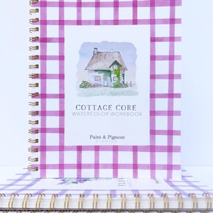 May include: A stack of watercolor workbooks with a pink and white gingham cover. The top book features a watercolor illustration of a cottage with a thatched roof and green ivy. The title of the book is "Cottage Core Watercolor Workbook" by Paint & Pigment Studios.