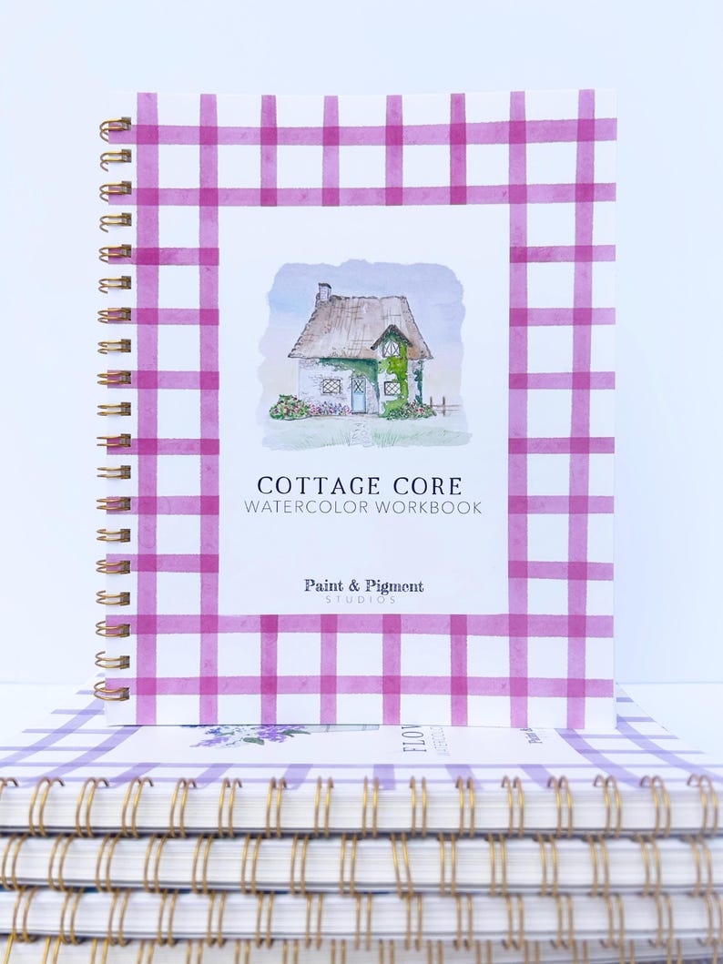 May include: A stack of watercolor workbooks with a pink and white gingham cover. The top book features a watercolor illustration of a cottage with a thatched roof and green ivy. The title of the book is "Cottage Core Watercolor Workbook" by Paint & Pigment Studios.