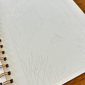 Puede incluir: Un cuaderno de bocetos con espiral con un boceto a l&aacute;piz de un paisaje. El boceto representa &aacute;rboles, agua y un horizonte distante. El papel es blanco y la encuadernaci&oacute;n es dorada.