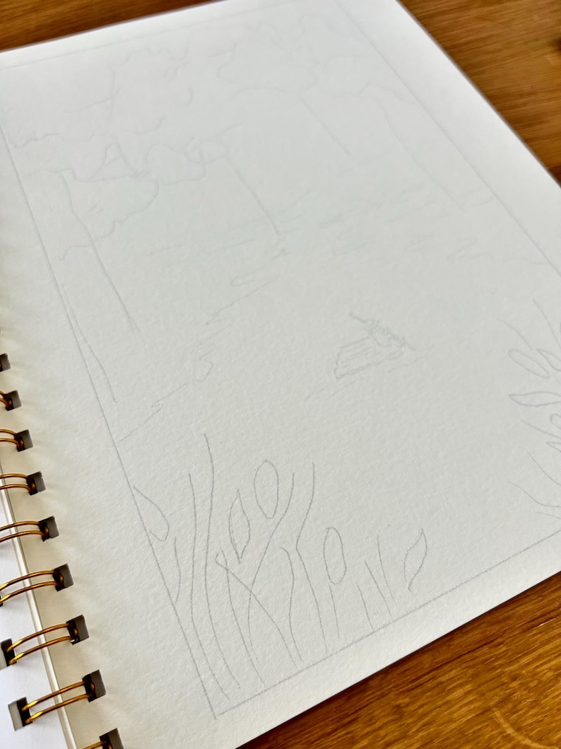 Puede incluir: Un cuaderno de bocetos con espiral con un boceto a l&aacute;piz de un paisaje. El boceto representa &aacute;rboles, agua y un horizonte distante. El papel es blanco y la encuadernaci&oacute;n es dorada.