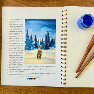 Puede incluir: Un diario de arte abierto con una acuarela de un perro en un paisaje nevado, acompa&ntilde;ado de un bote de pintura azul, dos pinceles y una paleta. El diario incluye instrucciones de pintura.