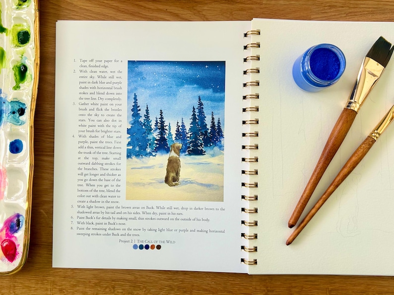 Puede incluir: Un diario de arte abierto con una acuarela de un perro en un paisaje nevado, acompa&ntilde;ado de un bote de pintura azul, dos pinceles y una paleta. El diario incluye instrucciones de pintura.