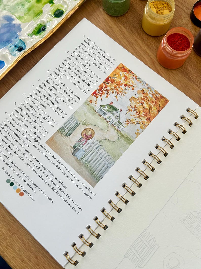 Puede incluir: Un libro de arte con espiral abierto en una acuarela de una casa con una valla blanca y follaje oto&ntilde;al. La p&aacute;gina incluye instrucciones y muestras de color. Se ven botes de pintura y una paleta.