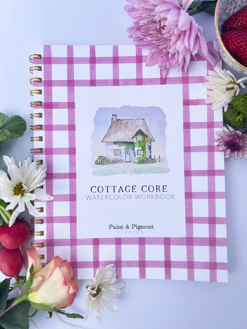 May include: A pink and white gingham notebook with a watercolor illustration of a cottage on the cover. The title of the notebook is "Cottage Core Watercolor Workbook" and the bottom of the cover reads "Paint & Pigment Studios".