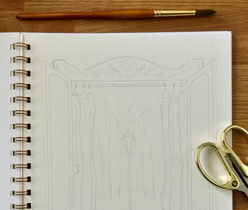 Puede incluir: Un cuaderno de espiral con un dibujo a l&aacute;piz de un armario. Un pincel y unas tijeras doradas est&aacute;n sobre la superficie de madera. El dibujo del armario tiene una parte superior decorativa con una cabeza de le&oacute;n y detalles florales.