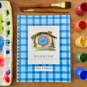 Puede incluir: Un cuaderno de acuarela con espiral titulado "Bookish" con una cubierta de cuadros azules. La portada presenta una ilustraci&oacute;n de una casa de hobbit. Pinceles, una paleta y botes de pigmento est&aacute;n colocados alrededor del cuaderno.