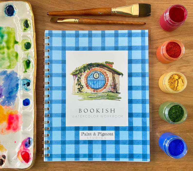 Puede incluir: Un cuaderno de acuarela con espiral titulado "Bookish" con una cubierta de cuadros azules. La portada presenta una ilustraci&oacute;n de una casa de hobbit. Pinceles, una paleta y botes de pigmento est&aacute;n colocados alrededor del cuaderno.