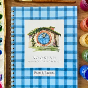 Puede incluir: Un cuaderno de acuarela con espiral y cubierta a cuadros azules. La portada presenta una ilustraci&oacute;n de acuarela de un agujero de hobbit con una puerta azul. El texto "BOOKISH WATERCOLOR WORKBOOK" est&aacute; impreso en la portada, junto con el logotipo "Paint & Pigment Studios".