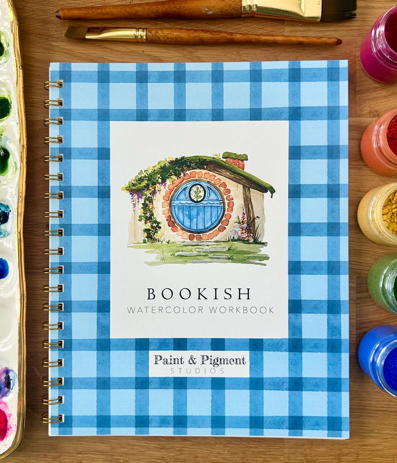 Puede incluir: Un cuaderno de acuarela con espiral y cubierta a cuadros azules. La portada presenta una ilustraci&oacute;n de acuarela de un agujero de hobbit con una puerta azul. El texto "BOOKISH WATERCOLOR WORKBOOK" est&aacute; impreso en la portada, junto con el logotipo "Paint & Pigment Studios".