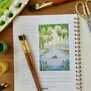 Puede incluir: Un diario de arte abierto con una pintura de acuarela de una escena de pantano, acompa&ntilde;ado de pinceles, pintura y una paleta. El texto del diario dice "Adventures of Huckleberry Finn."