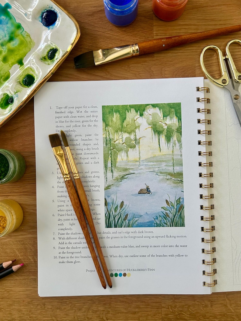 Puede incluir: Un diario de arte abierto con una pintura de acuarela de una escena de pantano, acompa&ntilde;ado de pinceles, pintura y una paleta. El texto del diario dice "Adventures of Huckleberry Finn."