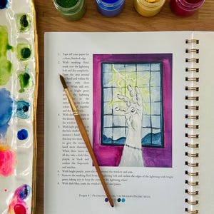 Puede incluir: Un diario de arte abierto con una ilustraci&oacute;n en acuarela de una mano que alcanza un rayo. Un pincel descansa sobre la p&aacute;gina. Frascos de pintura azul, amarilla, verde y roja est&aacute;n cerca, junto con una paleta de cer&aacute;mica.