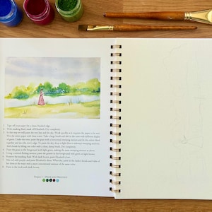 Puede incluir: Un cuaderno abierto con espiral con una pintura de acuarela de una persona con un vestido rosa junto a un lago. Tubos de pintura y pinceles sobre una superficie de madera. Una paleta de acuarela con colores mezclados a la izquierda.