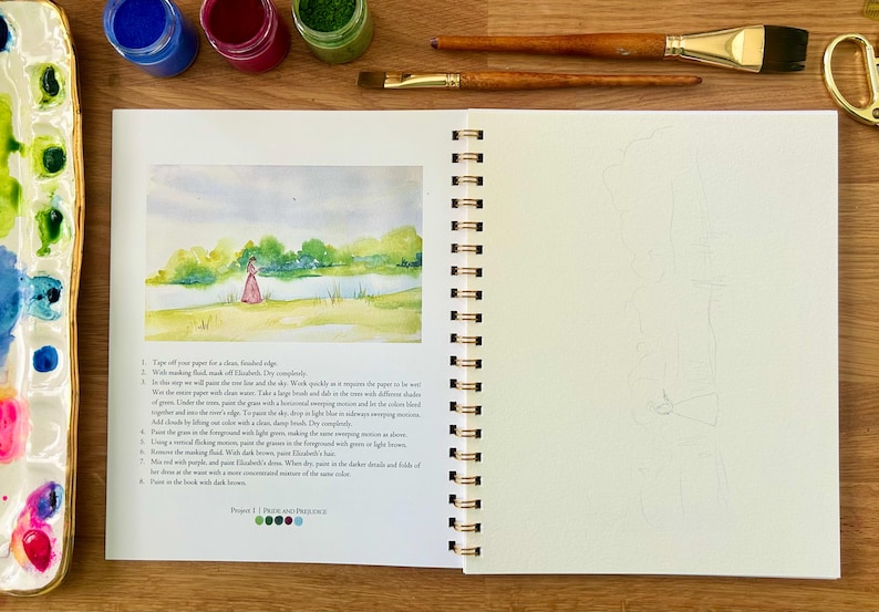 Puede incluir: Un cuaderno abierto con espiral con una pintura de acuarela de una persona con un vestido rosa junto a un lago. Tubos de pintura y pinceles sobre una superficie de madera. Una paleta de acuarela con colores mezclados a la izquierda.