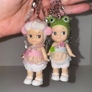 May include: Two small doll keychains with sparkly details. One doll is wearing a pink dress and a white hat with a sparkly pink and white design. The other doll is wearing a light blue dress and a green frog hat with sparkly green details.