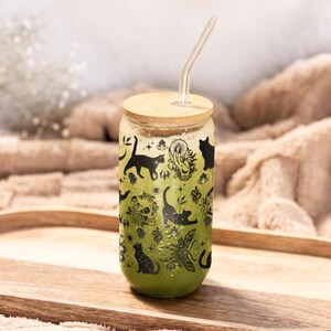 May include: A clear glass can-shaped tumbler with a bamboo lid and a glass straw. The tumbler is filled with a green liquid and decorated with black cat silhouettes and botanical illustrations. The tumbler sits on a wooden tray.