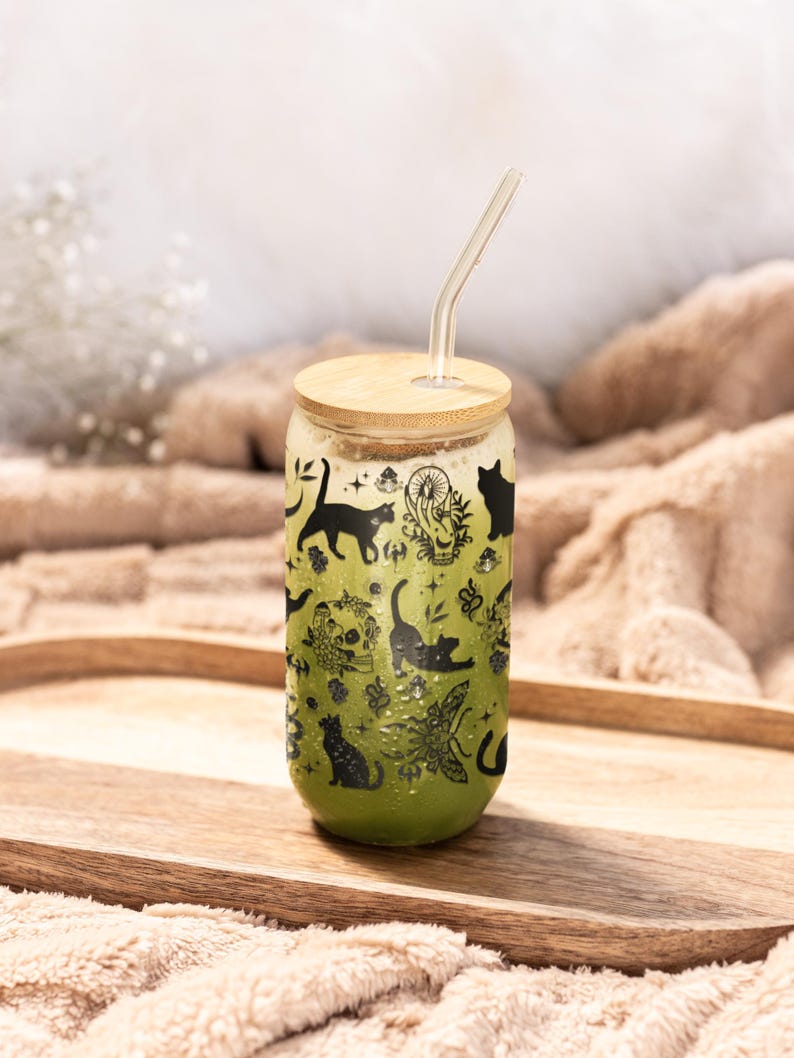 May include: A clear glass can-shaped tumbler with a bamboo lid and a glass straw. The tumbler is filled with a green liquid and decorated with black cat silhouettes and botanical illustrations. The tumbler sits on a wooden tray.