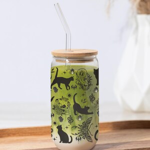 May include: A glass tumbler with a bamboo lid and a glass straw. The tumbler is filled with a green liquid and decorated with black cat silhouettes and other mystical illustrations. The tumbler sits on a wooden tray.