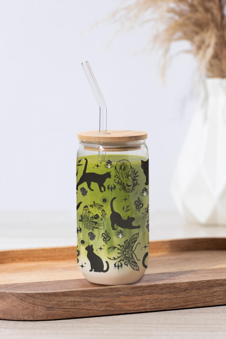 May include: A glass tumbler with a bamboo lid and a glass straw. The tumbler is filled with a green liquid and decorated with black cat silhouettes and other mystical illustrations. The tumbler sits on a wooden tray.
