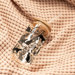May include: A clear glass tumbler with a bamboo lid and a clear straw. The tumbler is decorated with black cat silhouettes and other decorative elements. The tumbler is resting on a textured, peach-coloured fabric.