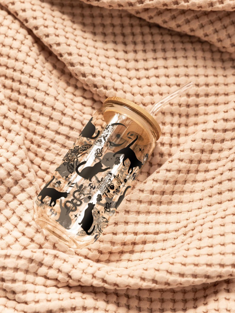 May include: A clear glass tumbler with a bamboo lid and a clear straw. The tumbler is decorated with black cat silhouettes and other decorative elements. The tumbler is resting on a textured, peach-coloured fabric.