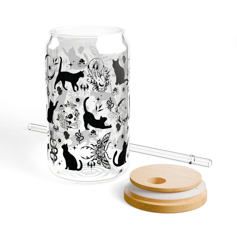 May include: A clear glass can-shaped drinking glass with a black cat and floral pattern. Includes a glass straw and a wooden lid. The glass is decorated with black cat silhouettes, skulls, and floral designs.