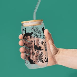 May include: A clear glass can-shaped tumbler with a bamboo lid and a clear straw. The tumbler is decorated with black cat silhouettes and other mystical illustrations. The tumbler is held by a hand against a teal background.