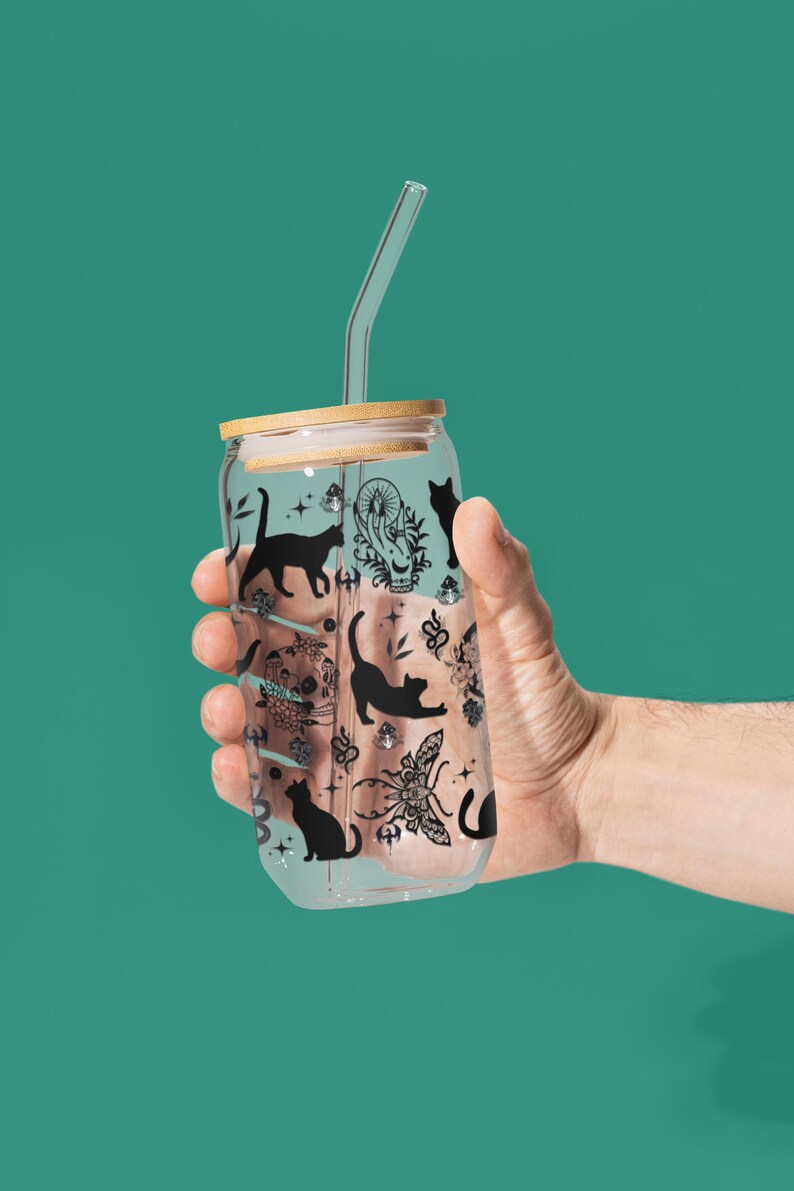 May include: A clear glass can-shaped tumbler with a bamboo lid and a clear straw. The tumbler is decorated with black cat silhouettes and other mystical illustrations. The tumbler is held by a hand against a teal background.