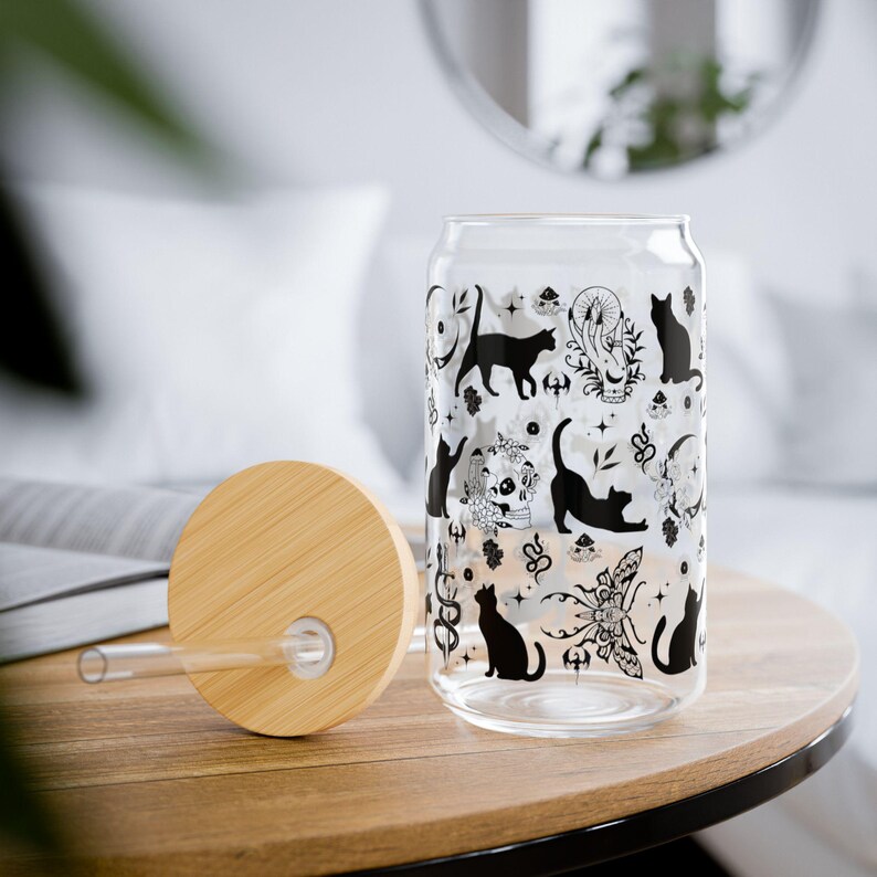May include: A clear glass can-shaped tumbler with a bamboo lid and straw. The glass is decorated with black cat silhouettes and occult-themed illustrations. The tumbler sits on a wooden surface.