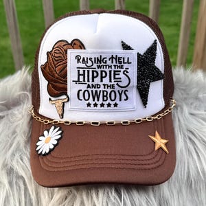 Puede incluir: Gorra de camionero marrón y blanca con la parte trasera de malla. La parte delantera presenta un parche con el texto "Raising Hell with the Hippies and the Cowboys", una rosa, una estrella y una margarita. Una cadena dorada y una estrella acentúan la visera.