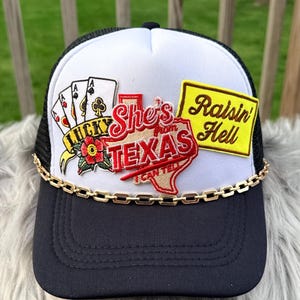 Op de afbeelding: Zwarte en witte truckerpet met een gouden ketting en patches. Patches zijn speelkaarten, een Texas-omtrek en de zinnen "Lucky", "She's from Texas I can tell" en "Raisin' Hell".