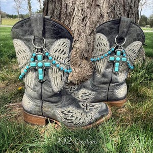 Puede incluir: Un par de botas de vaquero grises desgastadas con diseños bordados de alas y cruces. Cada bota presenta un colgante de cruz turquesa con detalles de cuentas a juego. Las botas están sobre un fondo natural.