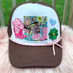 May include: A brown and white trucker hat with a pink beaded band. The hat features a patch with a pink cowboy hat and boots, flowers, and the text "Just a Small Town Girl". A blue heart and green flower are also on the hat.