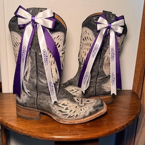 May include: A pair of distressed grey leather cowboy boots featuring intricate white embroidery and a cross design on the foot. Each boot is adorned with a decorative bow made of purple and white ribbons printed with the text Tarleton Texans.