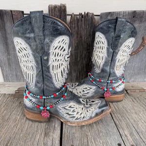 May include: A pair of distressed black cowboy boots with embroidered white angel wings. The boots are adorned with beaded anklets in red, turquoise, and silver, and a pink floral charm. The boots are displayed on a weathered wooden surface.