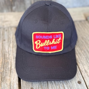 May include: Black baseball cap with a rectangular patch. The patch has a red background with yellow trim and the words "SOUNDS LIKE Bullshit TO ME" in white and red lettering. The cap is sitting on a wooden surface.