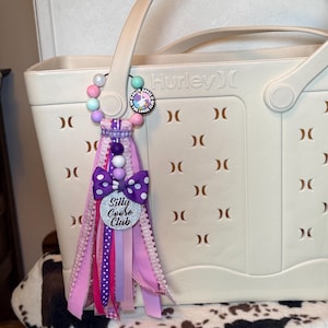 May include: A cream-colored Hurley tote bag with a decorative charm. The charm features a beaded strand, a purple polka dot bow, and a silver heart-shaped tag that reads "Silly Goose Club". The charm has pink and purple ribbons.