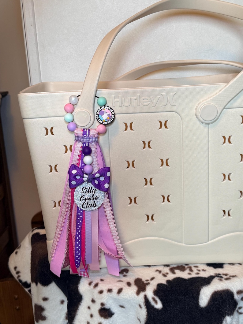 May include: A cream-colored Hurley tote bag with a decorative charm. The charm features a beaded strand, a purple polka dot bow, and a silver heart-shaped tag that reads "Silly Goose Club". The charm has pink and purple ribbons.