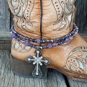 Puede incluir: Primer plano de una bota de vaquero de cuero marrón con intrincados diseños florales. Una cadena de bota de cuentas moradas con un colgante de cruz plateada cuelga de la bota. La cruz está adornada con cristales transparentes.
