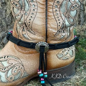 May include: A close-up of a tan leather cowboy boot with intricate floral and scrollwork designs. A black boot strap with a decorative silver buckle and dangling beads in shades of pink, teal, and silver adds a pop of color.
