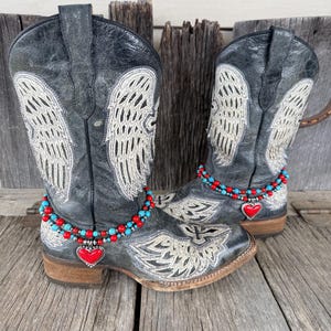 May include: A pair of distressed black leather cowboy boots with embroidered angel wings and decorative accents. The boots feature a square toe and a stacked heel. A beaded anklet with a red heart charm adorns each boot.