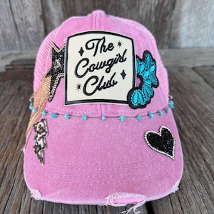 May include: Pink baseball cap with a distressed look. The cap has a patch that says "The Cowgirl Club" and is decorated with rhinestones, turquoise beads, and a black heart.