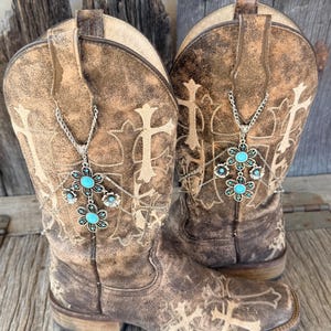 Puede incluir: Un par de botas de vaquero de cuero marrón desgastado con cruces bordadas y detalles florales. Cada bota tiene un collar plateado con colgantes de flores turquesas. Se exhiben sobre una superficie de madera envejecida.