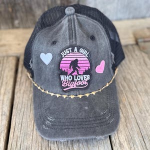 Op de afbeelding: Een zwarte truckerpet met een verwassen look en een mesh achterkant. De voorkant heeft een patch met een Bigfoot-silhouet, de tekst "JUST A GIRL WHO LOVES BIGFOOT" en twee hartvormige patches. Een gouden hartjesketting versiert de rand.