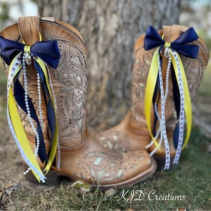May include: A pair of brown leather cowboy boots with decorative stitching. Each boot is adorned with a navy blue bow, yellow and navy ribbons, and a string of white pearls. The boots are decorated with football-themed ribbon.