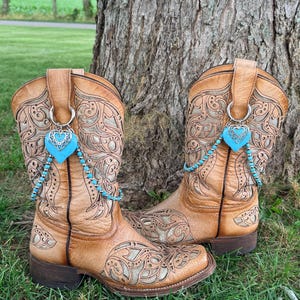 Può includere: Un paio di stivali da cowboy in pelle color cuoio con intricati motivi intagliati e uno strato metallico sottostante. Ogni stivale è decorato con un anello argentato che sostiene un ciondolo a forma di cuore turchese e una catenina.