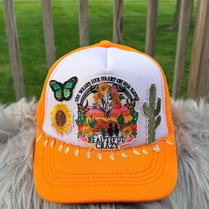 Puede incluir: Gorra de camionero naranja y blanca con la parte trasera de malla. La parte delantera presenta una colorida escena desértica con una calavera, girasoles y un cactus. También se ve el texto "She wears her heart on her sleeve" y "Beautiful Crazy".